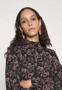 Blusa floral preta de mangas largas com decote e punhos com babados, apresentando padrões de flores cor-de-rosa e verdes. Brincos de gota coloridos e arrojados.