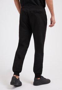 Schwarze Jogginghosen mit glatter Textur, elastischen Bündchen und einer Gesäßtasche. Kombiniert mit schwarzen Sneakers, die einen lässigen Look zeigen.