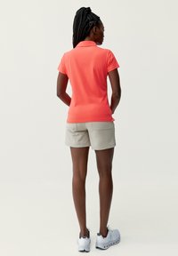 Koszulka polo z krótkim rękawem w kolorze koralowym, zestawiona z jasnobeżowymi szortami. Strój ma klasyczny krój, teksturowany materiał i brak widocznych wzorów.