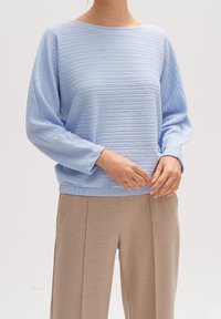 Jasnoniebieski sweter o fakturze, luźnym kroju i szerokich rękawach, zestawiony z beżowymi spodniami; ma dekolt marynarski i subtelne pionowe linie.