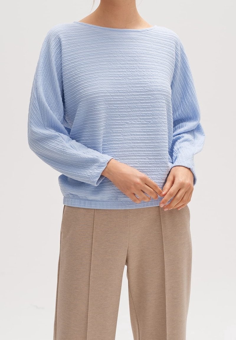 Jasnoniebieski sweter o fakturze, luźnym kroju i szerokich rękawach, zestawiony z beżowymi spodniami; ma dekolt marynarski i subtelne pionowe linie.