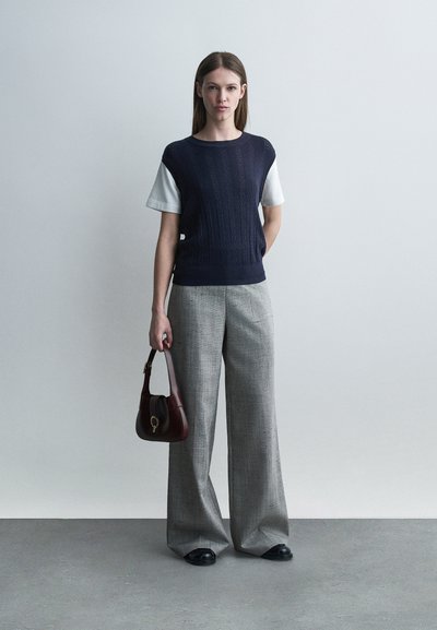 Γυναίκα που στέκεται μπροστά σε ένα απλό τοίχο, φορώντας μπλε σκούρο αμάνικο πουλόβερ πάνω από λευκά κοντομάνικα, γκρι παντελόνια με φαρδιά πόδια, μαύρα παπούτσια, κρατώντας καφέ τσάντα χειρός.