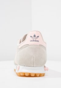 Světlé šedé semišové tenisky s růžovým akcentem na patě, vybavené texturovanou gumovou podrážkou a logem na zadní straně.