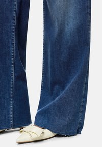 Wide-leg, dark blue denim jeans with frayed hem paired with white pointed-toe shoes featuring a bow detail.