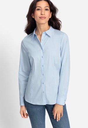 Frau, die ein hellblaues Hemd mit langen Ärmeln und blaue Jeans trägt, vor einem schlichten weißen Hintergrund.