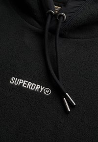 Sudadera con capucha de forro polar negro que presenta un logo bordado en plata "SUPERDRY" y cordones con puntas metálicas de marca. Tejido texturizado para mayor calidez.