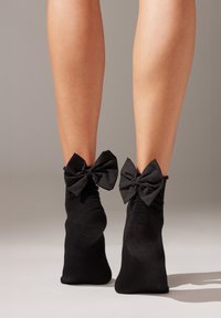 Schwarze Knöchelsocken mit großen Stoffschleifen am Bündchen. Glatte Textur mit klassischem Design, die auf nackten Beinen vor einem neutralen Hintergrund liegt.