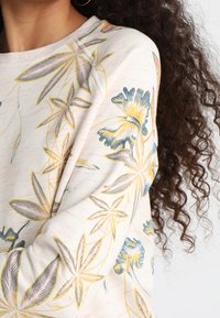 Beige Sweatshirt mit floristischem Muster, das lebendige blaue und gelbe Blumen zeigt. Weicher Stoff mit geripptem Halsausschnitt und strukturiertem Ärmeldesign.
