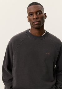 Άνδρας με κοντά μαλλιά που φοράει σκούρο γκρι φούτερ με το λογότυπο "LES DEUX", στέκεται μπροστά σε ένα απλό φωτεινό φόντο, κοιτάζοντας την κάμερα.