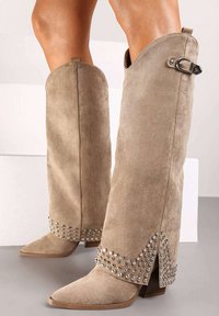 Bottes en suede beige jusqu'aux genoux avec des pointes, ornées de clous en argent au niveau des revers, dotées d'une boucle sur le côté pour l'ajustement.