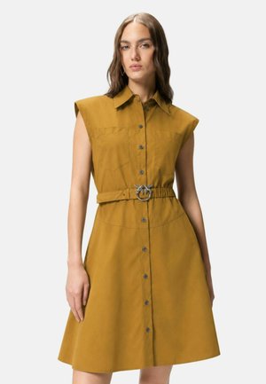 Femme portant une robe chemise sans manches couleur moutarde avec des boutons sur le devant et une ceinture assortie avec une boucle en argent ornée de deux oiseaux.