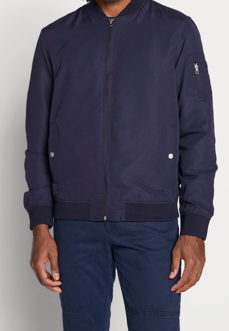 Granatowa bomberka wykonana z gładkiej tkaniny, z zamkiem błyskawicznym z przodu, ściągaczami przy mankietach, kieszeniami bocznymi oraz dodatkowymi kieszeniami zapinanymi na zamek na ramieniu.