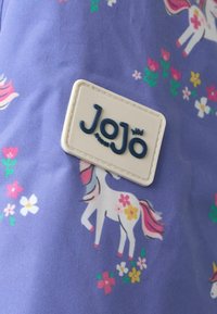 Lila Stoff mit einem verspielten Einhorn- und Blumenmuster. Ein rechteckiges weißes Etikett mit geprägt Text "JoJo" ist auffällig platziert.