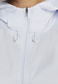 Primer plano de la cremallera de una chaqueta de color claro con cuerdas de tela y capucha, utilizada por una persona.