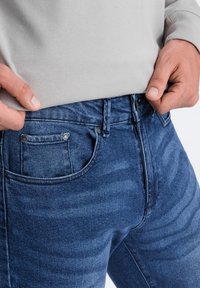 Tummaa sinisiä farkkuja, joissa on sileä pinta. Ominaisuuksiin kuuluu nappikiinnitys, useita taskuja ja hienovaraiset haalistumisviivat kankaassa.