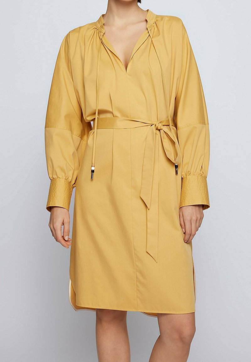 Robe jaune avec un col en V, un encolure froncée et de longues manches bouffantes. Elle présente une ceinture à nouer à la taille et des poignets texturés.