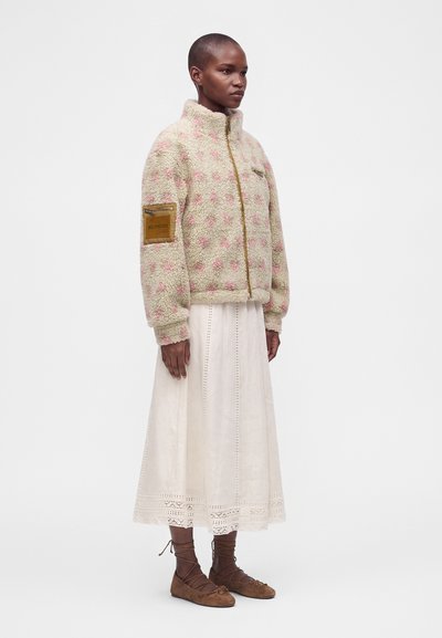 Femme portant une veste polaire beige et rose à motifs, une jupe blanche longueur cheville et des chaussures plates marron à lacets, debout sur un fond blanc uni.