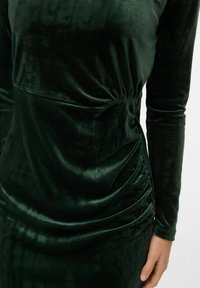 Zelena žametna obleka z dolgimi rokavi; vsebuje nabran detajl na pasu, gladko teksturo in prilegajoč silhuet.
