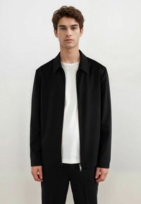 Svart zip-up jakke med kragehals, lange ermer og glatt tekstur, kombinert med en enkel hvit t-skjorte under.