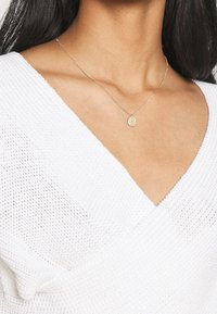 Gold pendant necklace on a fine chain, featuring a textured circular charm, worn with a white knitted V-neck top.