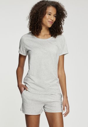 Young woman with curly hair wearing light gray short-sleeve shirt and matching shorts, standing with one hand in pocket, smiling.