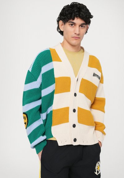 Cardigan tricoté coloré avec des rayures vertes, jaunes et bleues. Il présente un col en V, une fermeture à boutons et un emblème de smiley sur la manche.