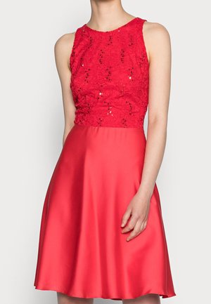 Rode mouwloze jurk met kanten en pailletten lijfje en een gladde satijnen uitlopende rok, gedragen door een persoon met één arm ontspannen langs het lichaam.