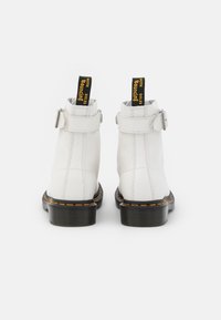 Białe skórzane botki za kostkę z paskiem z tyłu i klamrą, zaokrąglonym noskiem, żółtymi akcentami oraz czarną teksturowaną podeszwą. Na ściągaczu z tyłu znajduje się oznaczenie marki.