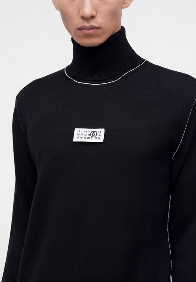 Homme portant un pull à col roulé noir avec des coutures blanches en contour et une étiquette rectangulaire blanche avec des chiffres sur la poitrine.