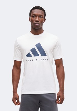 Ung mand iført en hvid T-shirt med stor mørkeblå Adidas-logo og "Real Madrid"-tekst, kombineret med mørkegrå shorts.