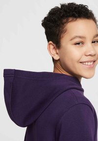 Sudadera con capucha morada de corte relajado, con capucha curvada y textura suave, vista desde el hombro izquierdo con una persona sonriente.