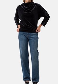 Zwarte fluwelen top met col en lange mouwen, gecombineerd met lichtblauwe jeans met rechte pijpen. De stof heeft een zachte textuur en een gladde afwerking.