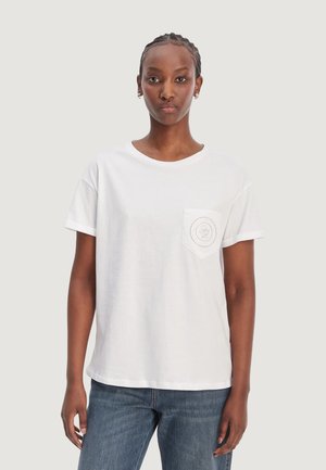 Femme aux cheveux tressés portant un T-shirt blanc à manches courtes avec un logo sur la poche poitrine et un jean bleu, debout devant un fond clair.