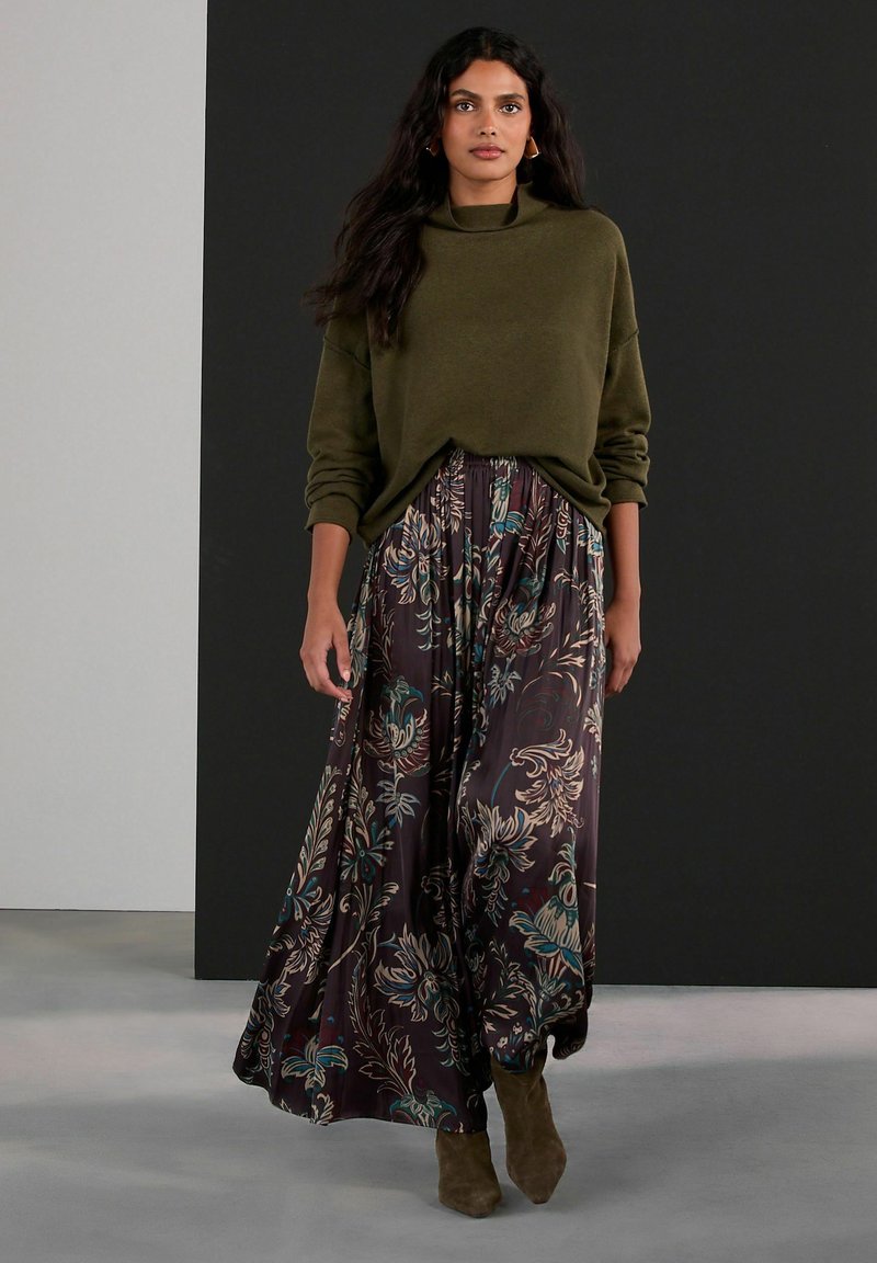 Olivově zelený svetr s vysokým límcem; splývavá maxi sukně s tmavým květinovým vzorem s tyrkysovými a krémovými akcenty; taupe kotníkové boty.