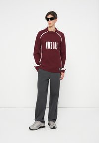 Bordo pulover s kapuco z logotipom "NIKE AIR", belimi detajli in sivimi trenirkami. Model nosi črne sončne očala in sive superge. Preprost ozadje.