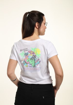 T-shirt en coton blanc avec un imprimé graphique coloré au dos, comprenant le texte "Salutations de" et "LES AVENGERS !", le tout sur un fond de palmiers et de soleil.