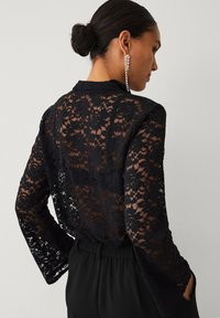 Schwarze Spitzenbluse mit langen Ärmeln, floralen Mustern und lockerem Schnitt. Dazu schwarze Hosen mit elastischem Bund.