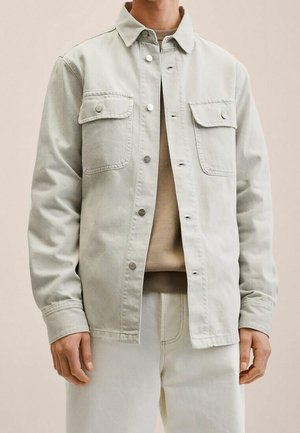 Man draagt een lichtbeige jas met knopen en twee klepzakken aan de voorkant over een tankleurig shirt en gebroken witte broek, staand tegen een effen achtergrond.