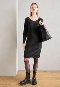 Tmavě šedé žebrované pletené šaty s výstřihem do V, dlouhými rukávy a přiléhavým střihem, doplněné černými kotníkovými botami a černou kabelkou přes rameno.