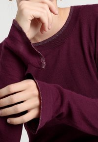 Ręce korygują rękawy osoby noszącej purpurowy sweter z długim rękawem, z lekko postrzępionymi mankietami i okrągłym dekoltem.
