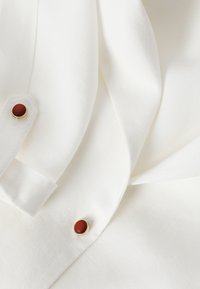 Tissu blanc avec deux boutons ronds marron sertis d’or sur un vêtement plié, mettant en valeur la texture et les détails des boutons.