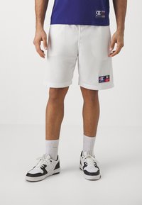 Pantalones cortos deportivos de malla blanca con dobladillo enrollado, que cuentan con un parche de logo. Combinados con zapatillas negras y blancas y calcetines blancos de caña corta.
