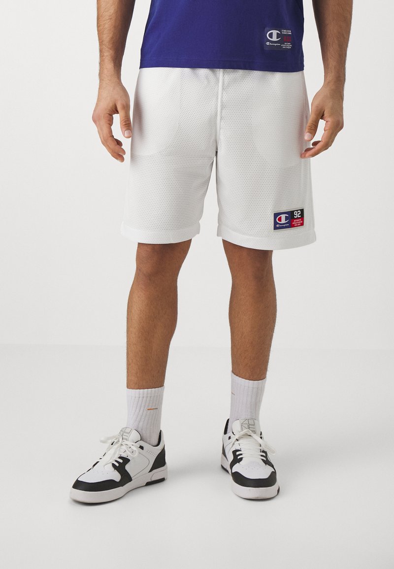 Pantalones cortos deportivos de malla blanca con dobladillo enrollado, que cuentan con un parche de logo. Combinados con zapatillas negras y blancas y calcetines blancos de caña corta.