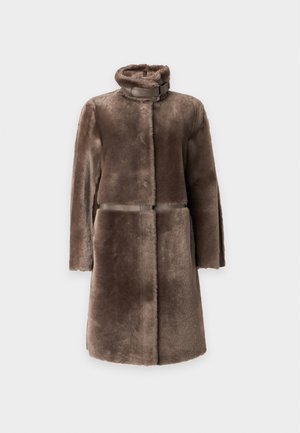 Femme portant un long manteau en fourrure marron, un pantalon beige à jambes larges et des ballerines noires pointues, debout devant un fond clair uni.