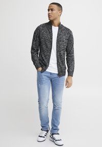 Suéter de cremallera en negro y gris jaspeado, camiseta blanca debajo, jeans claros desgastados y zapatillas con detalles en azul marino.