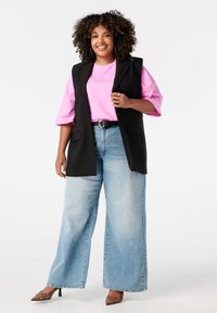 Glunderende vrouw met krullend haar, gekleed in een roze shirt, een zwarte mouwloze blazer, wijde spijkerbroeken, panterprint hakken en zilveren creolen.