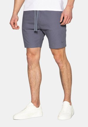 Homme portant un short gris à cordon, des baskets blanches et une chemise blanche, debout devant un fond clair uni.