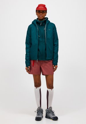 Person står iført en teal hættetrøje, mørkerøde shorts, rød kasket, hvide knælange sokker med stribe og grå vandresko mod en neutral baggrund.