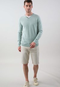 Un pull en tricot bleu clair avec un col en V et trois boutons décoratifs au col, porté avec un short beige clair retroussé et des baskets blanches.