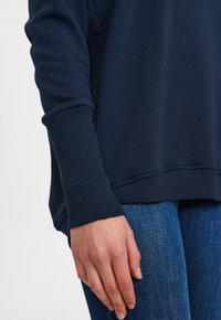 Sötétkék pulóver bordázott mandzsettával, puha tapintással és laza szabással, kék farmernadrággal párosítva. A mandzsetta részletére fókuszálva.